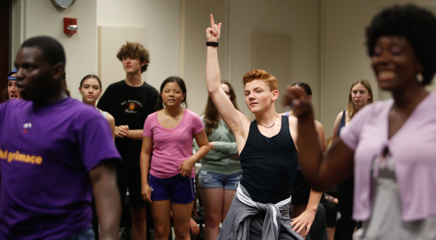 A group of dancers are in various poses. The focus is on a young red-headed boy with his finger in the air like a 70s dancer.