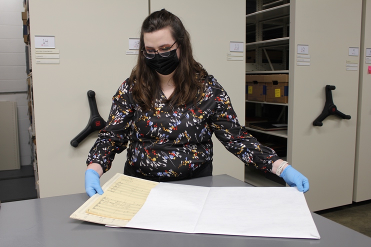 Breyanne pulls a musical arrangement from a white envelope. She wears a face mask and floral top.