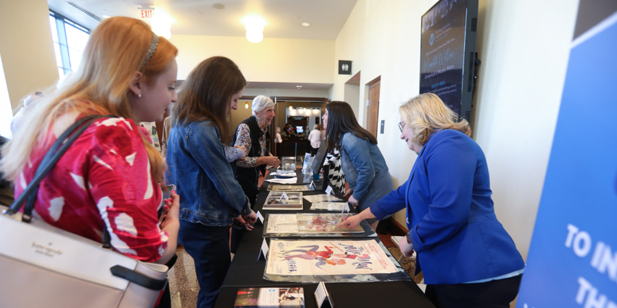 Two women stand behind a table and point to paper artifacts while women on the other side of the table look down at the artifacts.